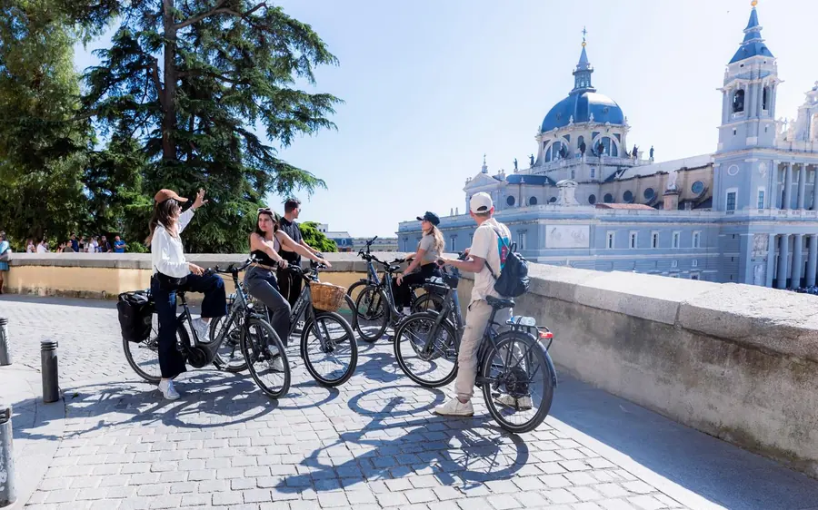 Riders pausing with their bikes on a walkway, the dome and towers of the Almudena Cathedral behind them