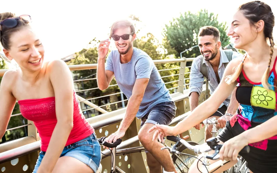 Four riders side by side on a bridge in the sunshine, laughing as they pedal