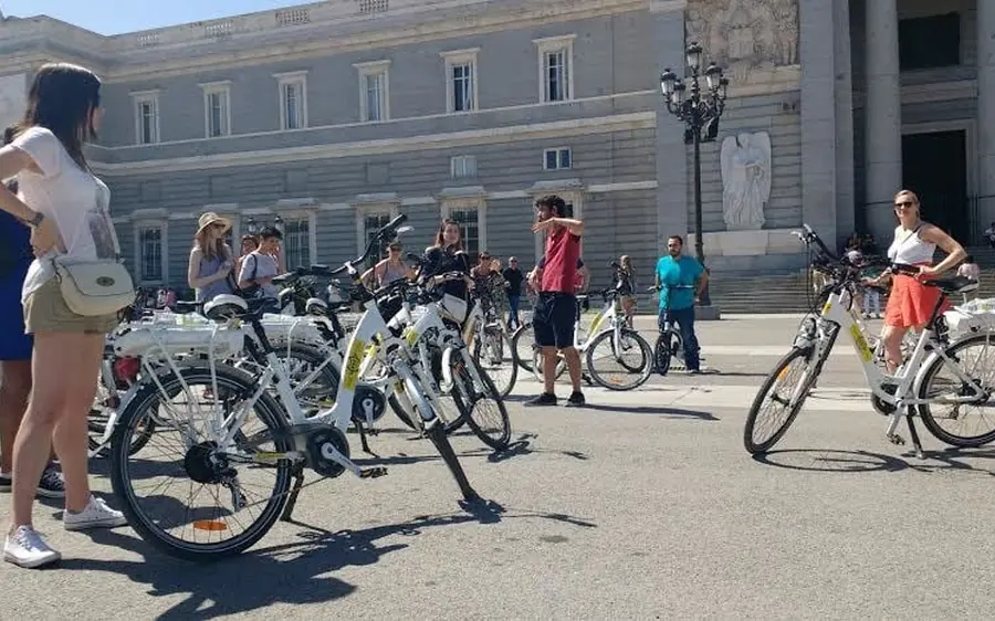 A row of white electric bikes and their riders in front of the long facade of the Royal Palace