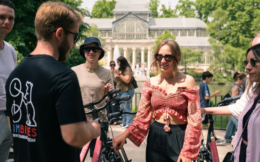 A guide in a black T-shirt talking to riders beside their bikes in front of a glass pavilion in the park