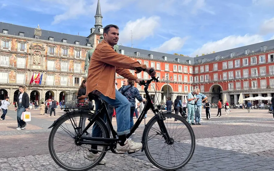 A man riding a black bike across the Plaza Mayor, the red-brick arcades behind him