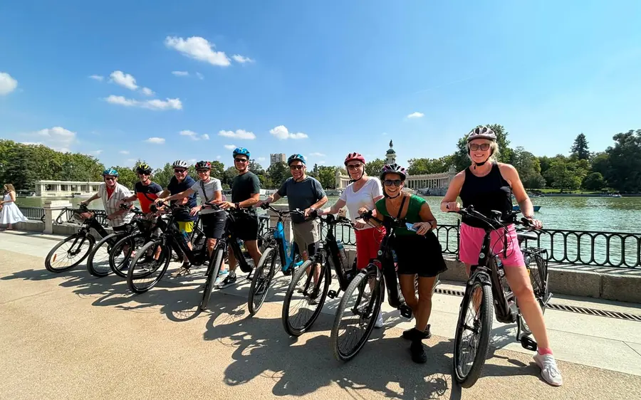 A group of riders in helmets with their bikes on a riverside walkway, one of them raising a hand to the camera